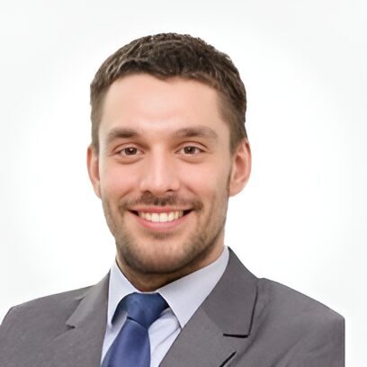 Man with short brown hair and beard, wearing a gray suit, white shirt, and blue tie, smiling against a light background.